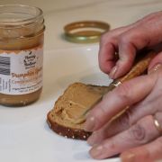 Hands spread pumpkin spice creamed honey on a slice of bread; jar labeled 'Pumpkin Spice Creamed Honey' sits nearby on a counter.