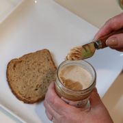 Person spreads pumpkin spice creamed honey from a jar onto a slice of whole-grain bread on a white plate as a kitchen scene unfolds