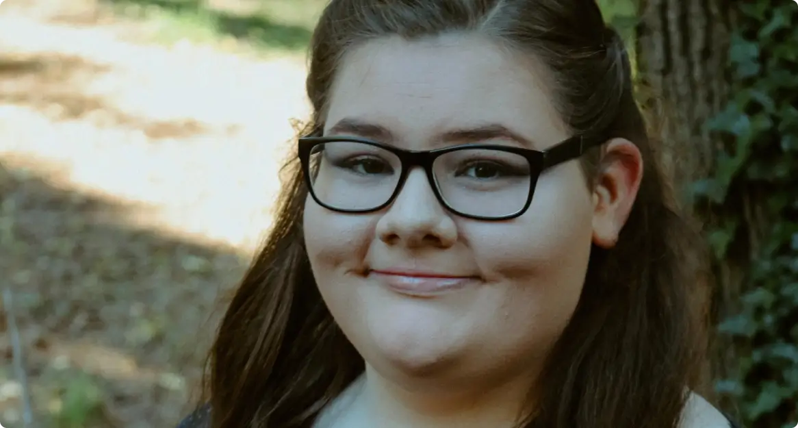Young woman with dark hair and black glasses smiling outdoors with trees in the background