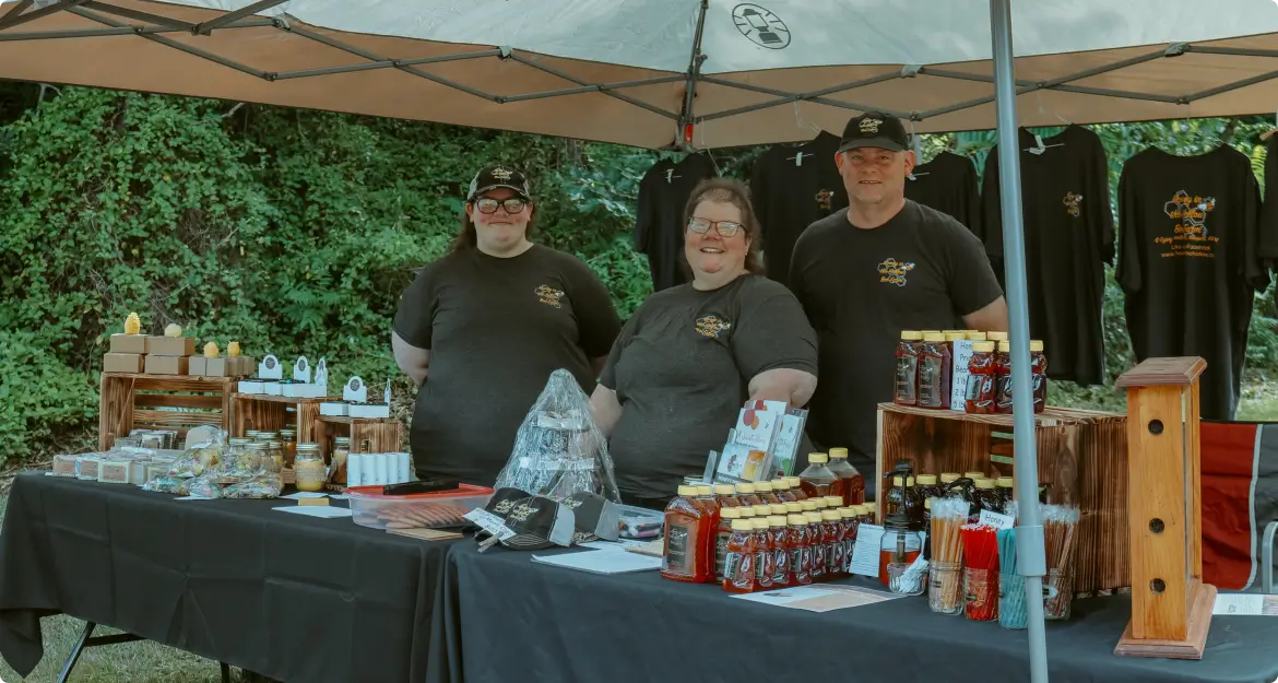 Three vendors stand behind a long table of honey jars and crafts at an outdoor market stall under a canopy