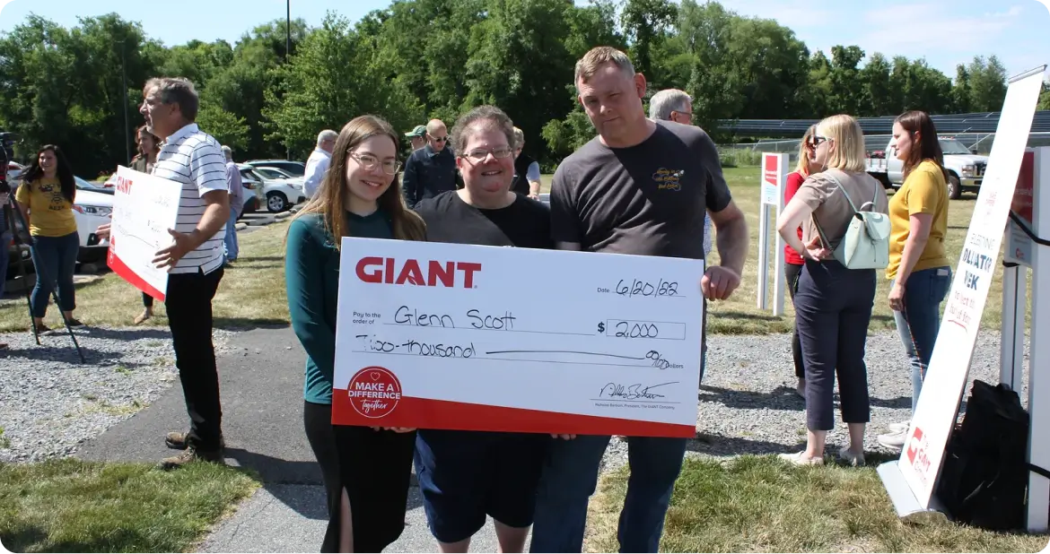 Group of people posing outdoors with a large GIANT charity check for $ made out to Glenn Scott smiling at a donation event on a sunny day