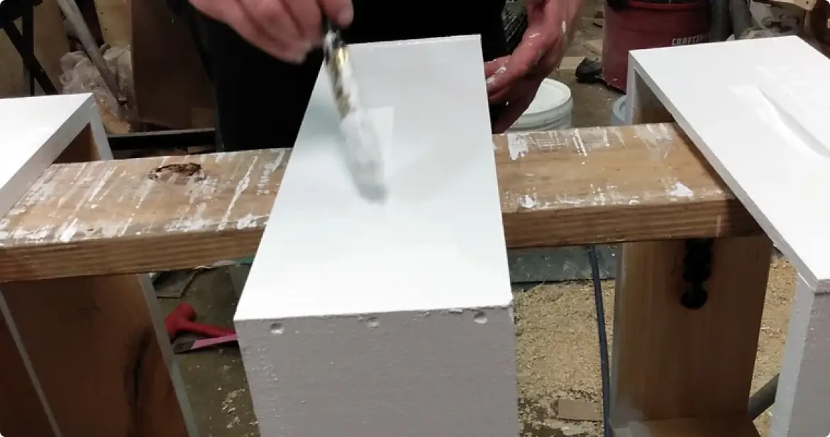 Person applying white paint to the top of a rectangular white wooden board on a workbench in a workshop