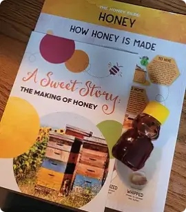 Two honey focused books on a wooden surface titled A Sweet Story The Making of Honey and Honey How Honey Is Made with a small honey jar beside them
