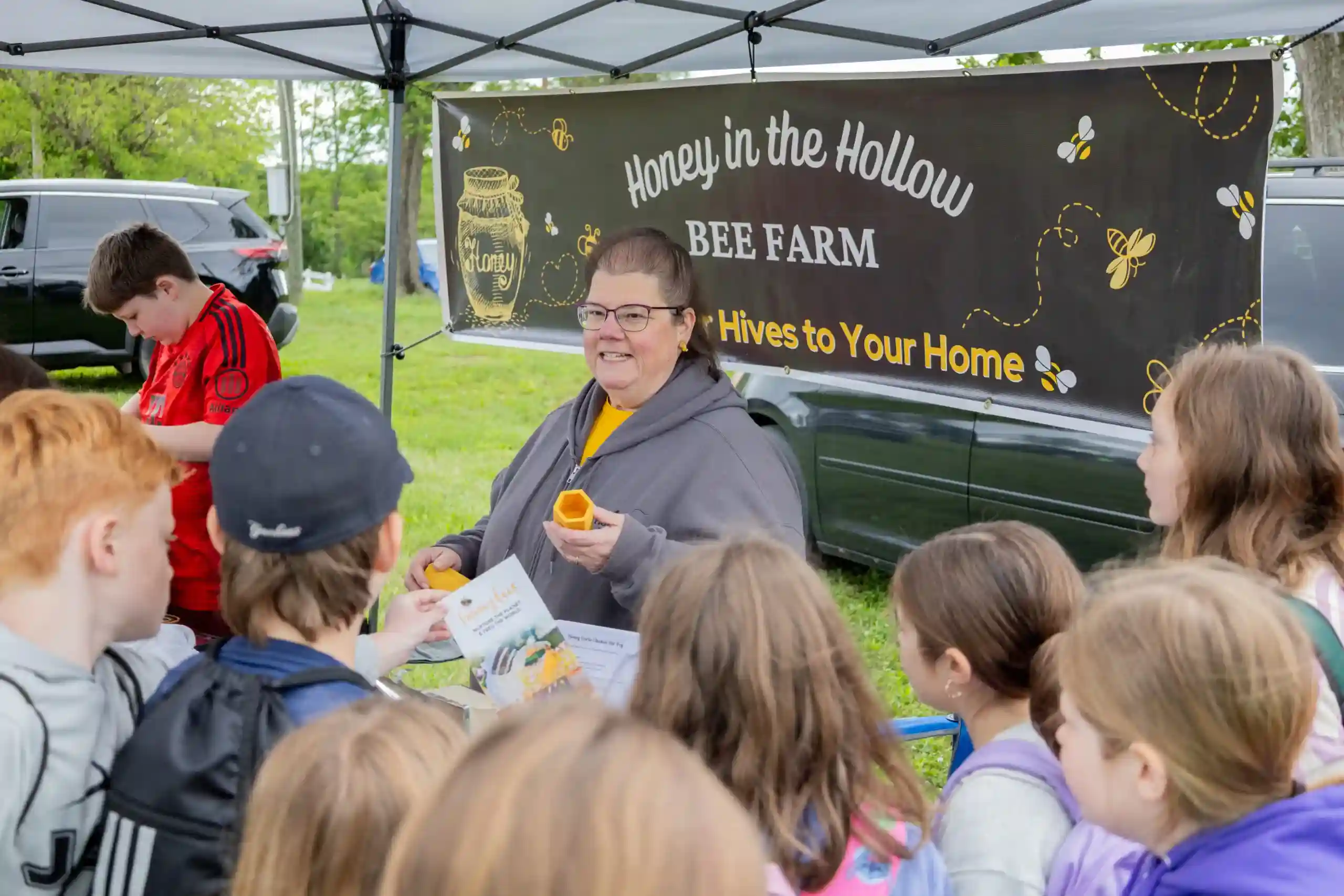 Children gather around a woman under a canopy at a bee farm booth handing out flyers as a banner reads Honey in the Hollow Bee Farm and Hives to Your Home