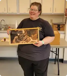 Smiling woman in a dark T shirt holding a wooden framed photo of a dog in a room with cabinets and a table behind her