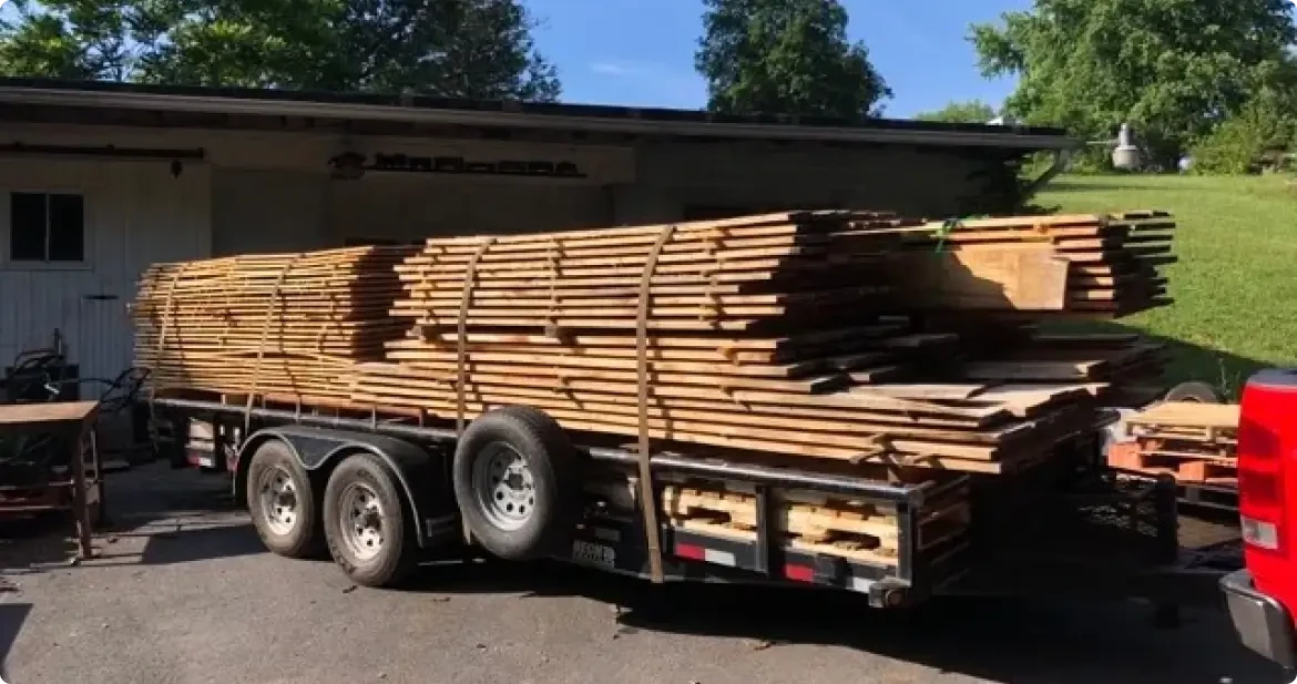 Trailer loaded with stacked lumber tied down parked beside a shed on a sunny day