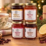 Four jars of infused honey (Lemon, Rose, Hibiscus, Cinnamon) arranged on a wooden table with a cup of tea and holiday decorations in the background.